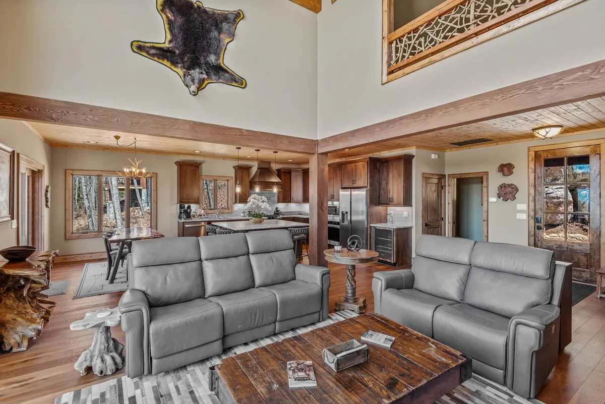 The image captures an open-concept interior space, showing plush leather sofas arranged around a wooden coffee table, with the kitchen area visible in the background. Large windows offer expansive mountain views, flooding the room with natural light and visually connecting the interior to the outdoors. The overall mood is warm and welcoming, perfect for gatherings.