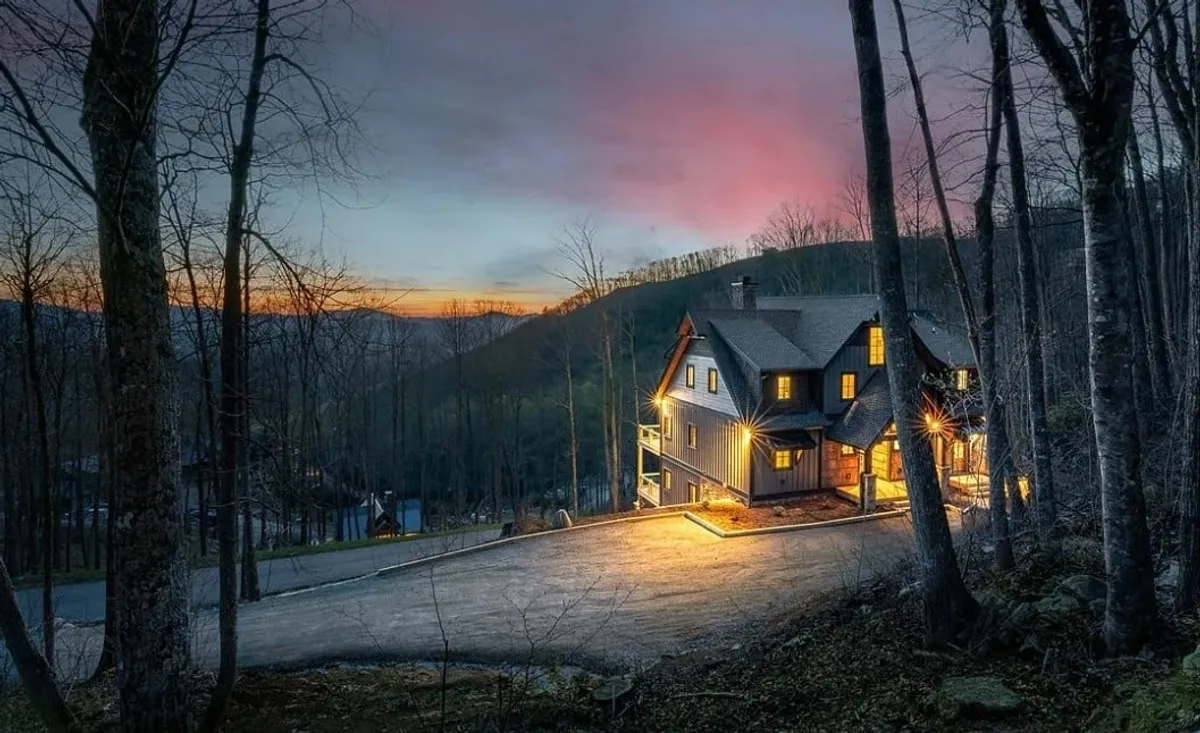 The image captures the house at sunset, illuminated with warm light, set against a backdrop of surrounding trees and an open sky. The scene suggests evening gatherings, nature walks, and a peaceful connection to the mountain environment—a perfect setting for returning from outdoor adventures with pets.