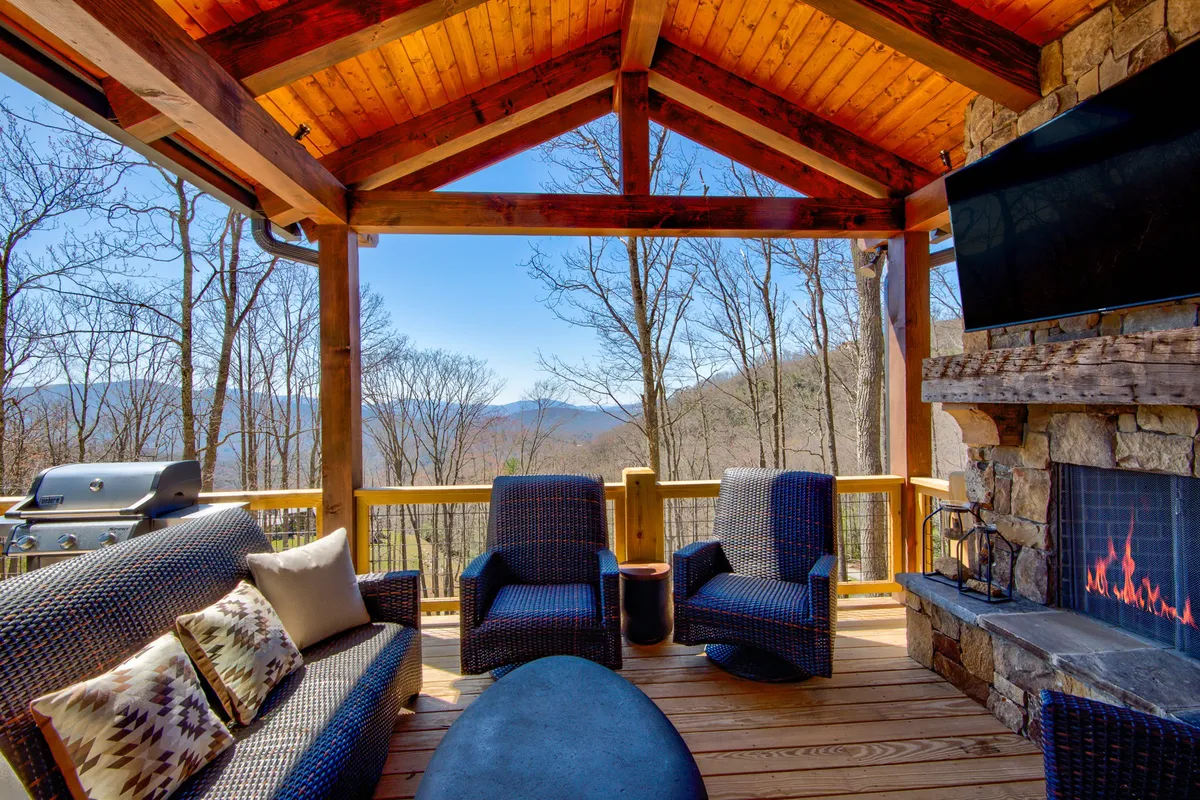 This exterior image showcases an inviting outdoor seating area on a spacious deck, featuring a stone fireplace, modern grill, flat-screen TV, and stunning mountain views. The arrangement invites relaxation and social gatherings, capturing the essence of al fresco living.