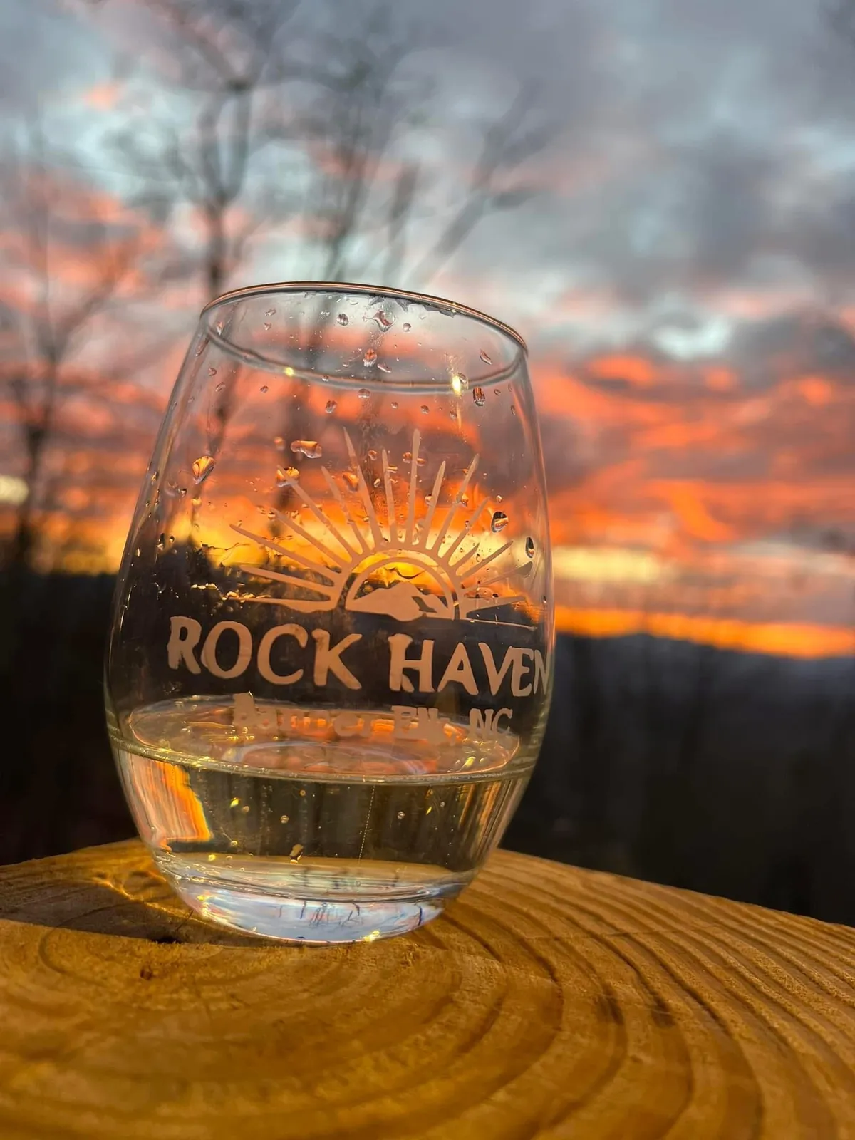 A close-up of a glass of wine set on a wooden surface outdoors at sunset, with soft golden light illuminating the scene. The background hints at natural surroundings and the tranquil ambiance of enjoying a drink outside, possibly with a loved one.