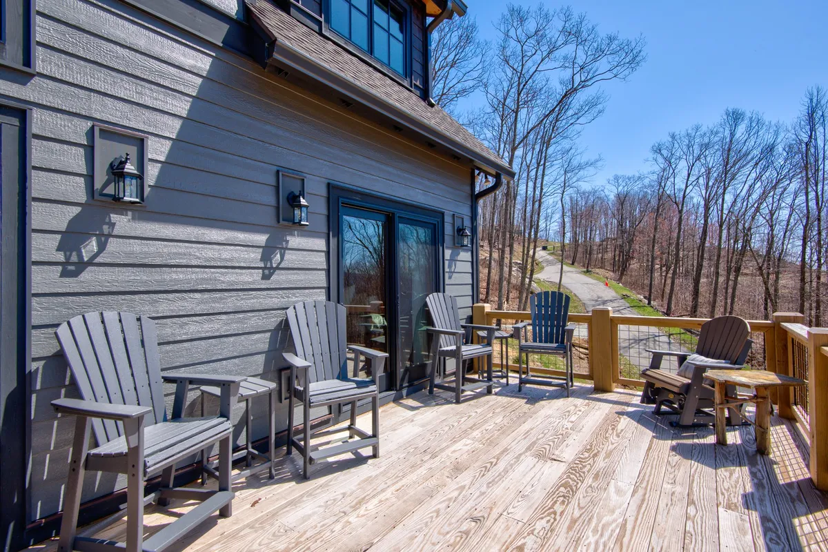 A wooden deck with Adirondack chairs overlooks a wooded area, with a pathway visible in the background among dense trees. The inviting seating arrangement suggests a place for relaxing after a day of adventure or gathering with friends around the fire pit. The connection to the outdoors is central, with the deck serving as an interface between home comforts and wilderness.