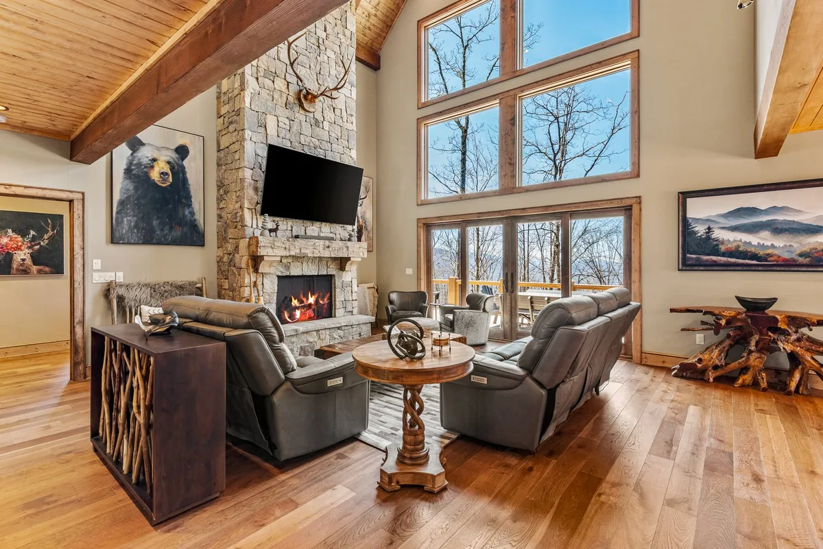 A spacious living room showcases a grand, floor-to-ceiling stone fireplace as its centerpiece, surrounded by rustic wooden furniture and tall windows framing a lush forest view. Artwork adds a personal touch while the inviting furniture arrangement encourages both relaxation and entertaining.