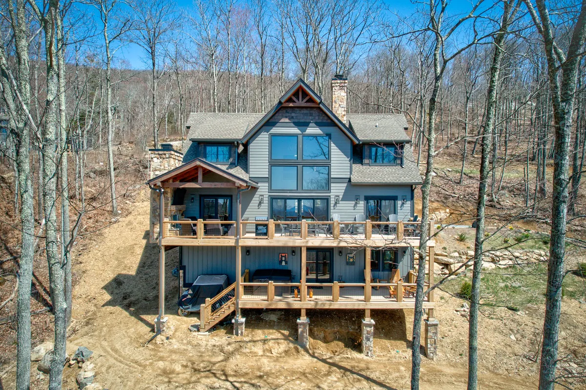 A wide view of a large wooden deck with expansive windows and a stone chimney, surrounded by trees. The deck is spacious, inviting, and perfectly suited for outdoor gatherings, with sunset light warming the scene and suggesting possibilities for group socializing, grill nights, or cozy evenings by the fire pit.