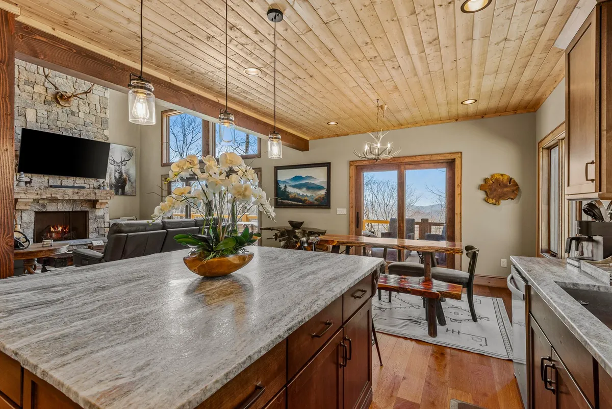 The image displays an open-plan space where a kitchen island with a marble countertop, a stone fireplace, a large dining table, and expansive windows all coexist. Natural light pours in, highlighting the harmony between cooking, dining, and relaxing areas.