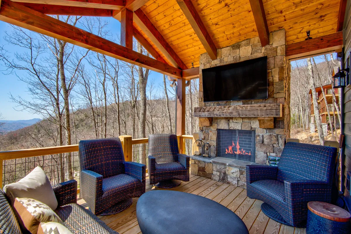 A cozy outdoor living area featuring a fireplace, comfortable seating, and a television, all under a wooden ceiling. The arrangement is ideal for relaxing with friends, enjoying a movie, or gathering around the fire as the sun sets, blending indoor comfort with an outdoor setting.